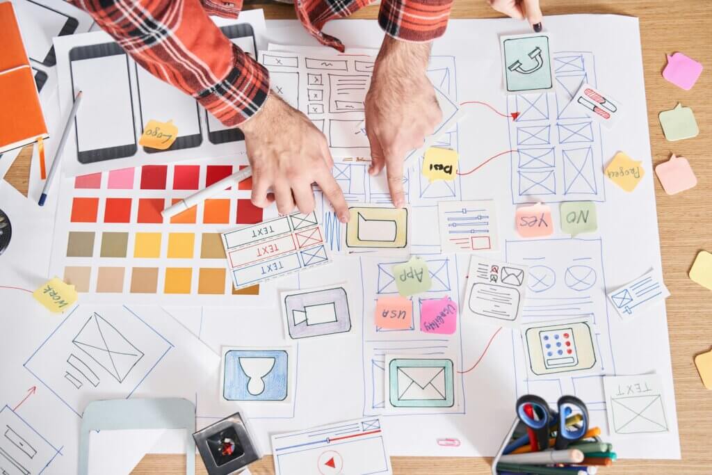 Ein Tisch ist voll mit Skizzen und Papier-Prototypen für eine digitale Benutzeroberfläche. Mehrere Hände sortieren gezeichnete UI-Elemente wie Buttons, Icons und Layouts. Daneben liegen Farbfächer, Post-its und Stifte verstreut, wodurch der Eindruck eines aktiven, kreativen Workshop-Prozesses entsteht.